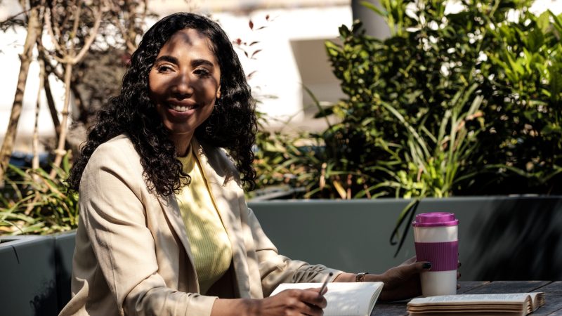 Smiling woman on a break holding cup of coffee and studying from a book. She is also writing on a notebook. Some plants on the background. No more technology concept.
